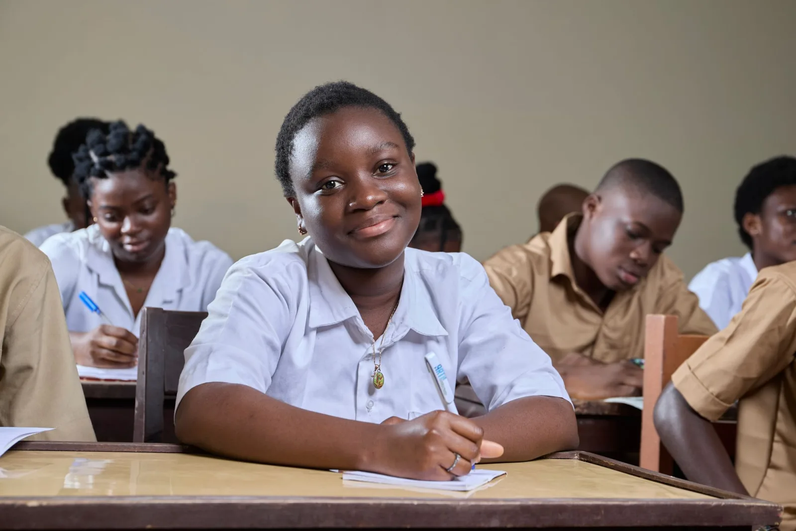 A student in a white uniform smiling at her desk in a Nigerian classroom. Photo by Mickael Ange Konan on Pexels.